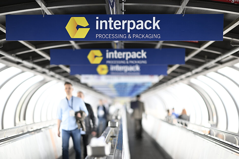 Mensen lopen op een rolband onder meerdere blauwe borden met het gele Interpack-logo en de tekst 'Interpack PROCESSING & PACKAGING' in een tunnelachtige constructie.